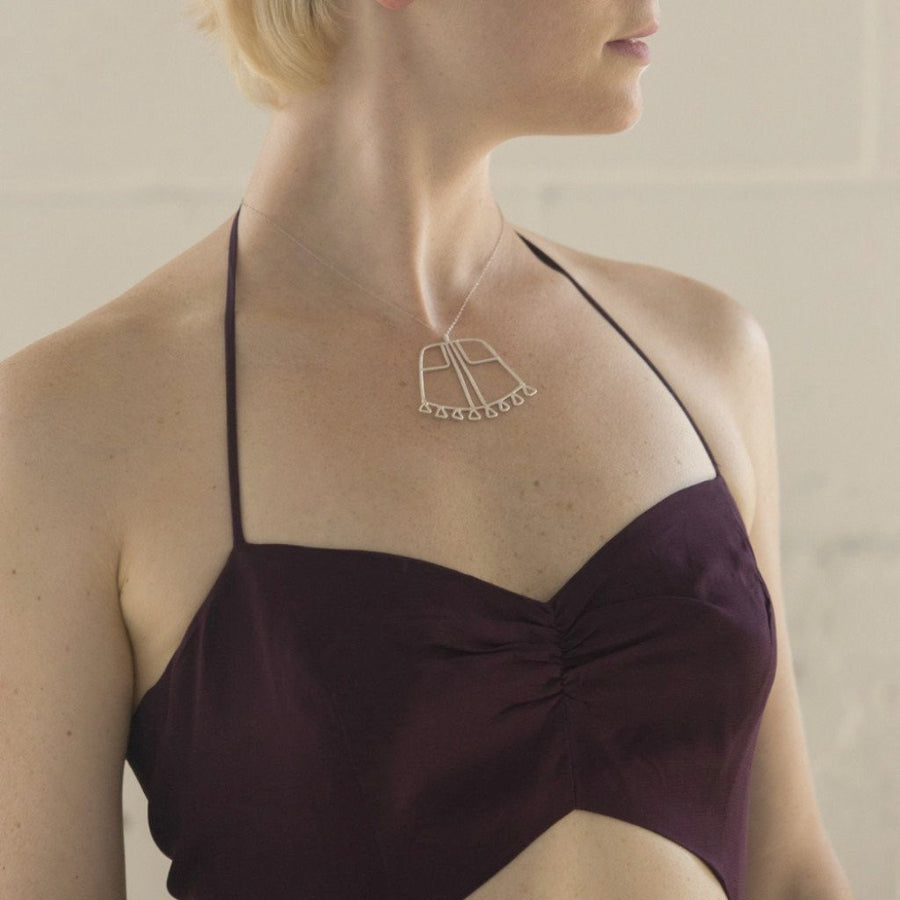 Model wears sterling silver Sky Phaebl necklace. Trapezoid pendant features linear filigree detail and triangular fixed fringe along the curved base. Pendant hangs on delicate chain and lays just below the clavicle.   